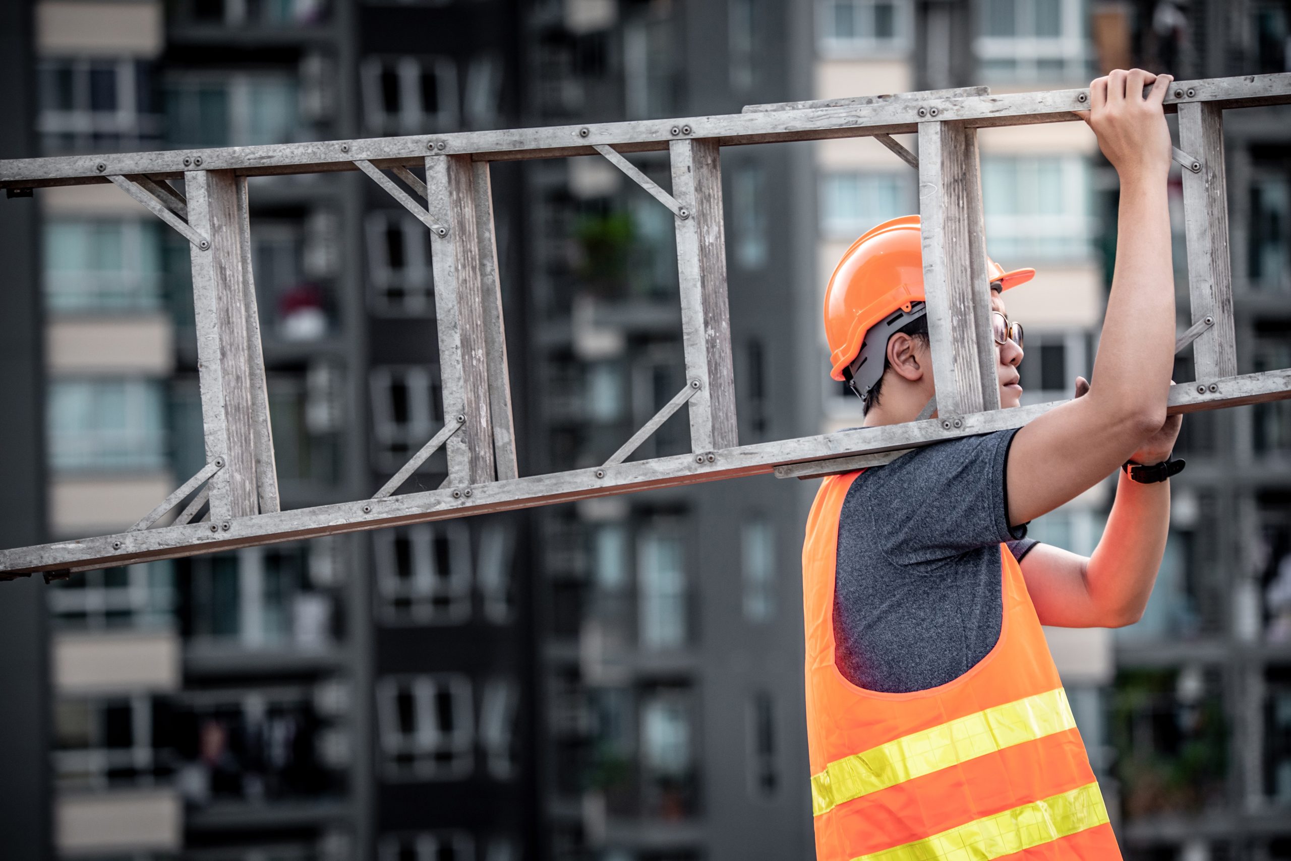 maintenance worker with orange safety helmet and vest carrying aluminium step ladder at construction site. Civil engineering, Architecture builder and building service concepts
